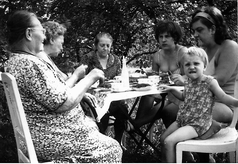 Schwarz-weißes Foto aus der Kindheit von Heike König. Fünf Frauen sitzen an einem Tisch im Garten, vor ihnen stehen Teller mit Kuchen. Ganz rechts zieht ein Kind, Heike König, eine Grimasse und schaut in die Kamera